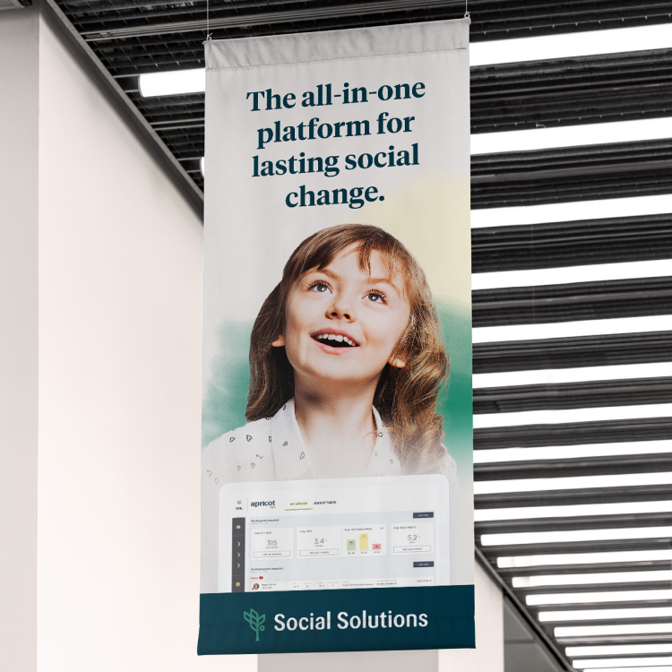 a mockup of a banner hanging in an office with the tagline "the all-in-one platform for lasting social change" above a photo of a young girl looking upwards with an expression of wonder and a picture of a tablet with the apricot software pulled up