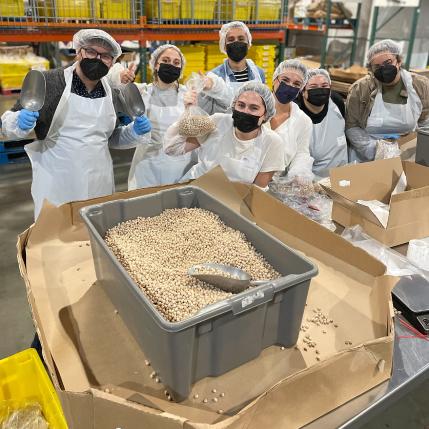 Five people wearing hair nets and aprons stand by a bin of food at a food pantry