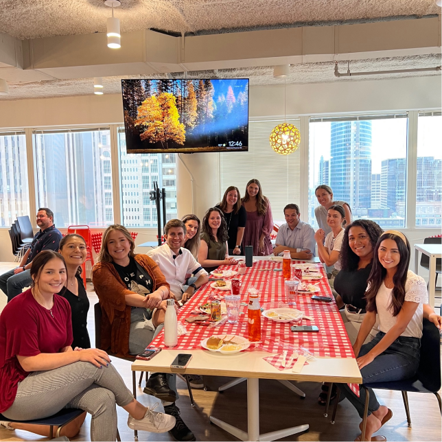 A group of people smiling while sitting at a table with a red checkered table cloth and enjoying a pot luck