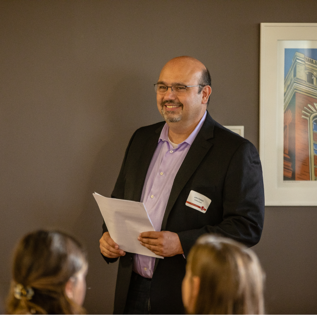 A man smiling in front of a brown wall while holding a piece of paper looking off to the left