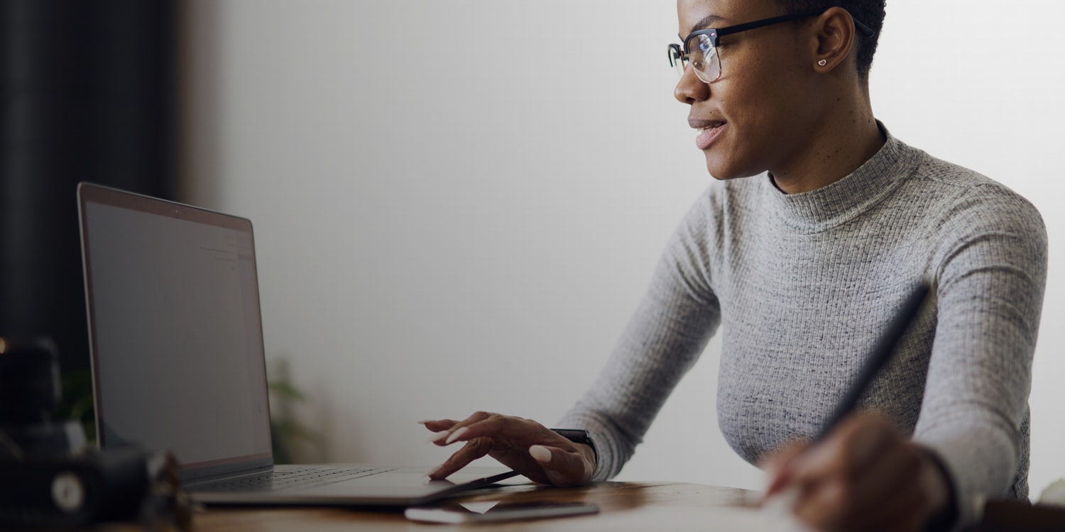 close-up photo of a woman sitting at a desk looking at a laptop and holding a pen