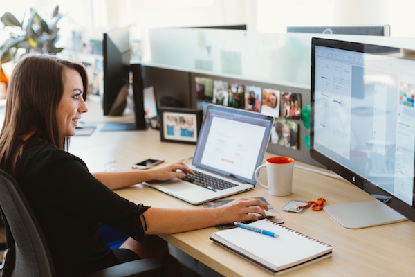 Woman in black shirt working at desk in an office with a laptop and monitor on her desk