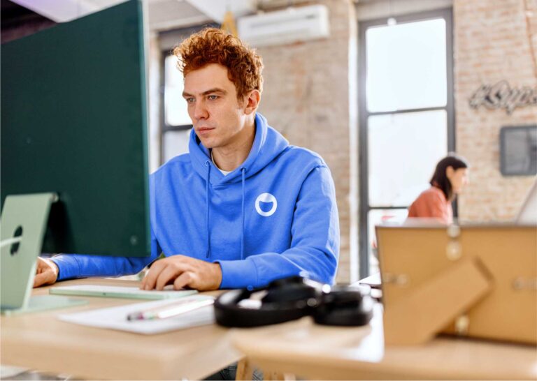 a man in a light blue hoodie with the ebbo logo typing at a desktop computer in front of an exposed brick wall