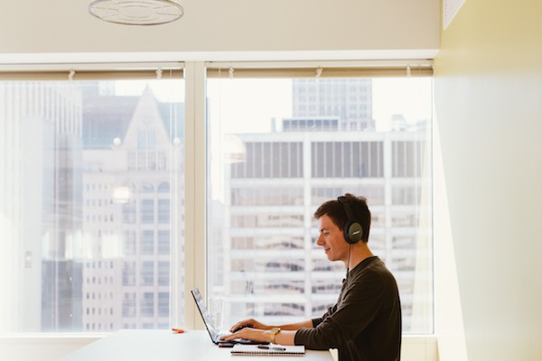 Man with headphones on standing at a table working on his laptop 