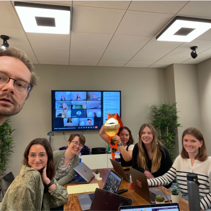 Group of employees sitting together a table while also on Zoom with other team members, displayed on a monitor on the wall