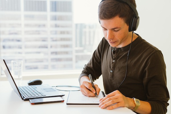Man in brown shirt wearing headphones and writing in notebook