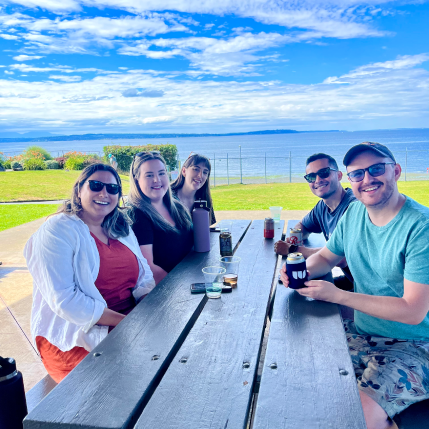 Five people sitting at a park table surrounded by grass and water. 