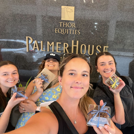 Four women holding chocolate treats in front of a building that says "Palmer House." 