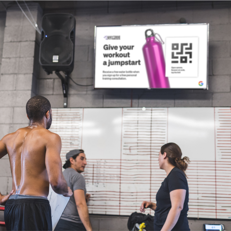 Three people standing and working out in a gym with a TV screen displaying a purple water bottle and text that reads: "Give your workout a jumpstart"