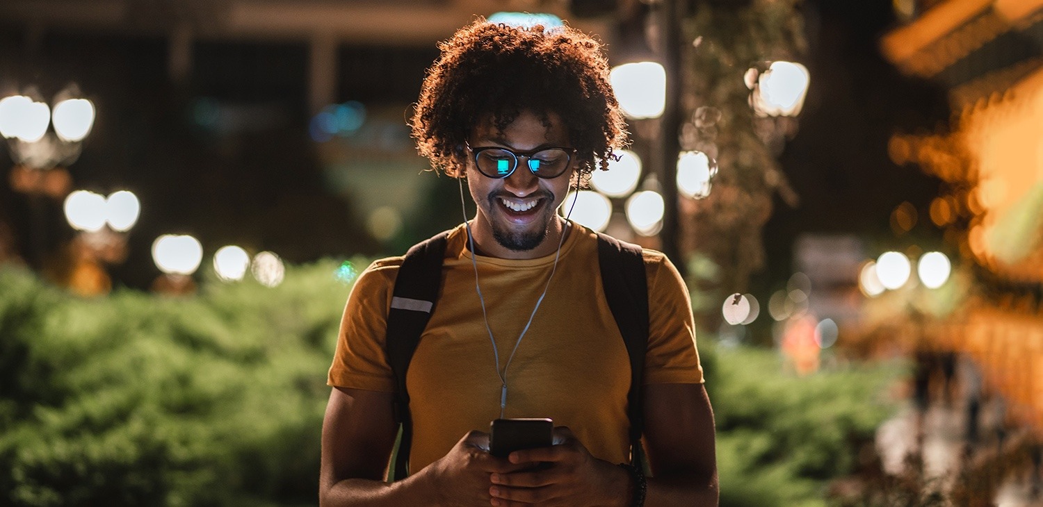 a man in a yellow shirt with a backpack looking down at his smartphone and smiling