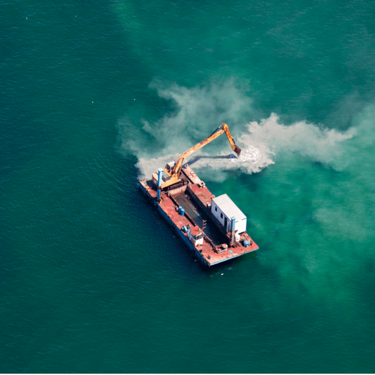 An aerial view of an industrial barge with a yellow excavator parked on its deck, anchored in a calm blue ocean