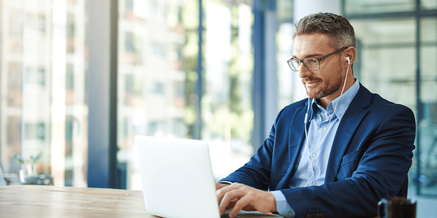 man in a navy suit with earbuds in typing on a laptop in front of office windows