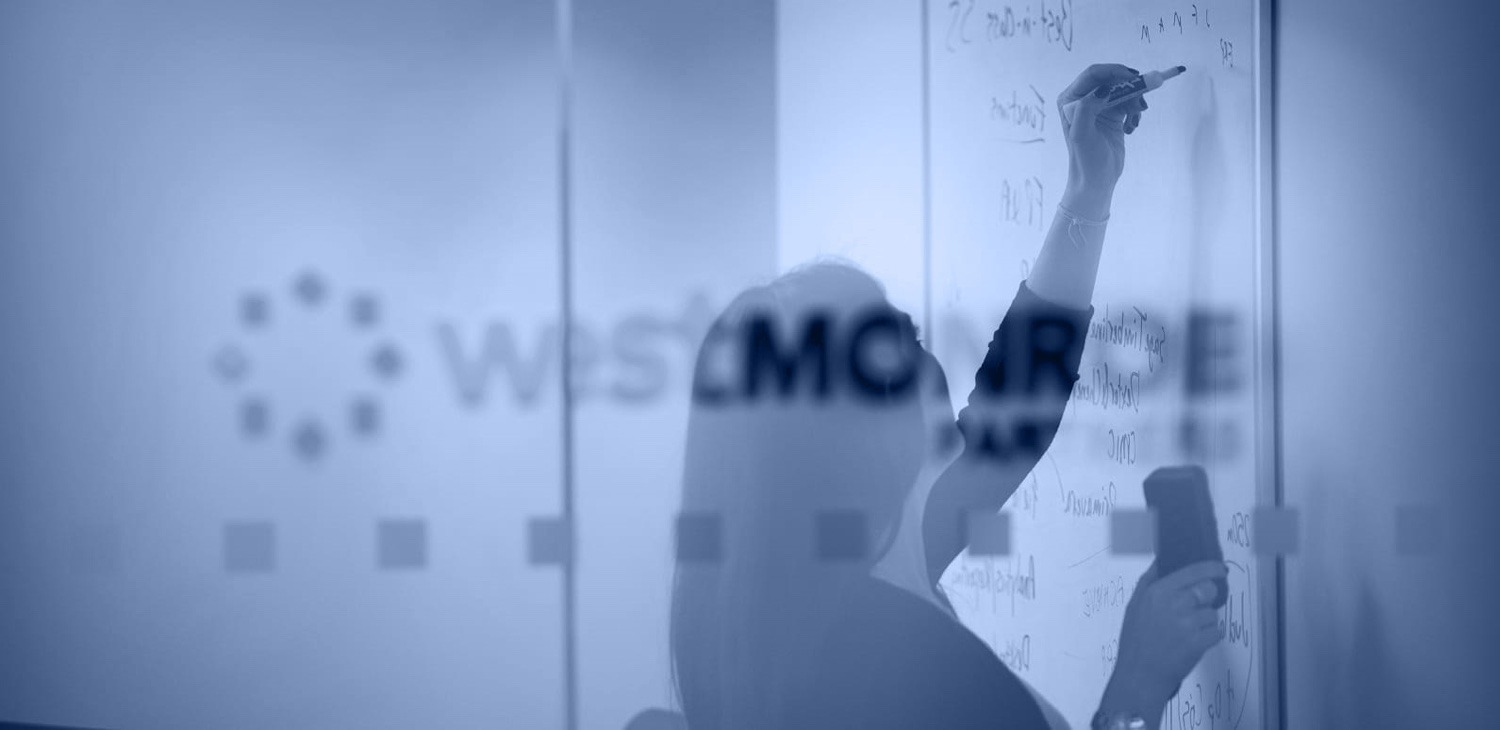 photo of a woman writing on a white board, shot through the glass door of a conference room with the West Monroe Partners logo