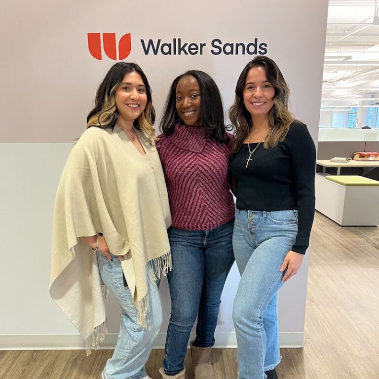 Three women standing in Chicago office in front of wall with Walker Sands logo