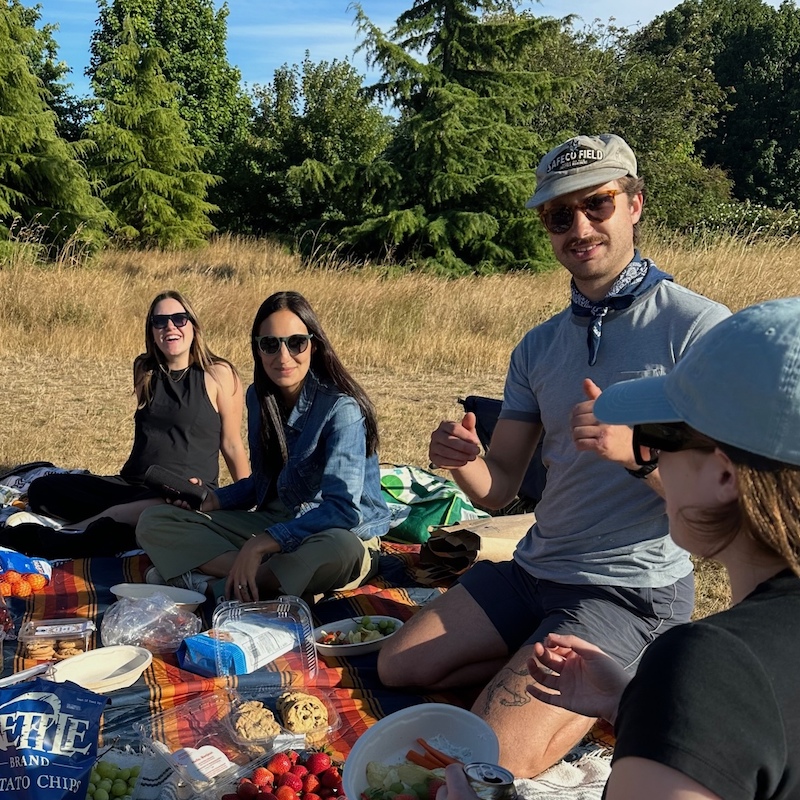 For people sitting on a picnic blanket having a picnic with snacks with trees in the background