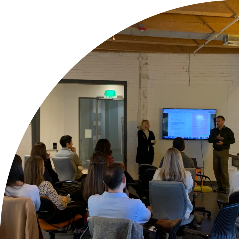 two people presenting powerpoint slides to an audience of nine or ten people in a loftlike room with whitewashed brick walls